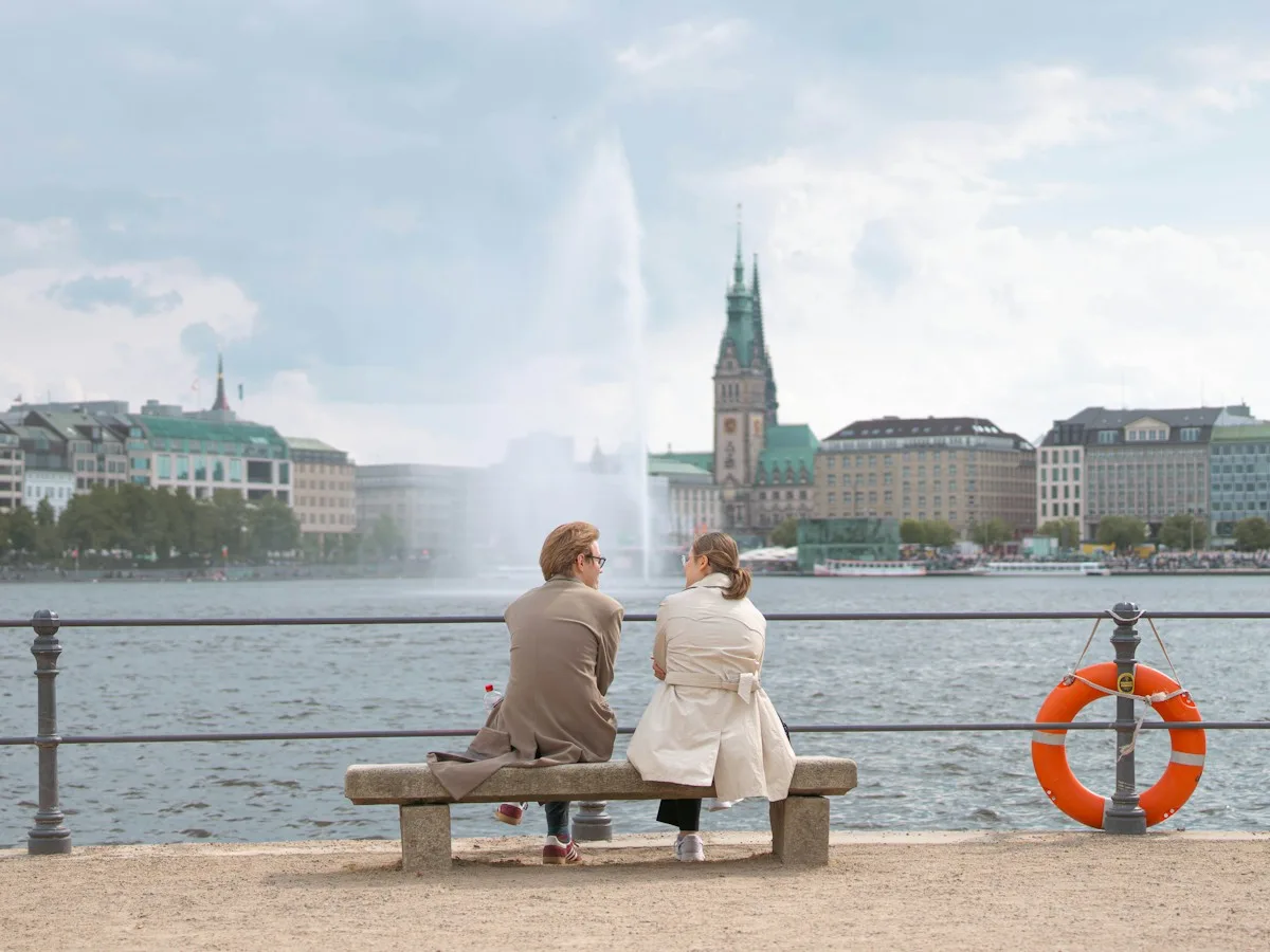 Two people sitting on a bench by the Alster, looking across the water at Hamburg