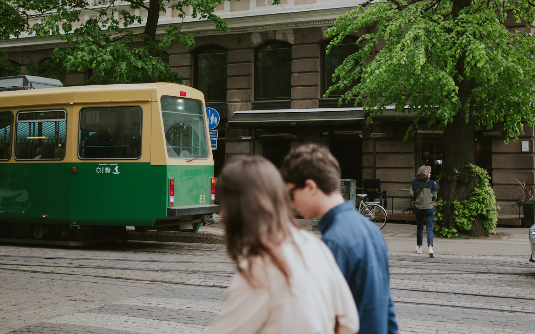 A couple walking past a green tram on a leafy Helsinki street