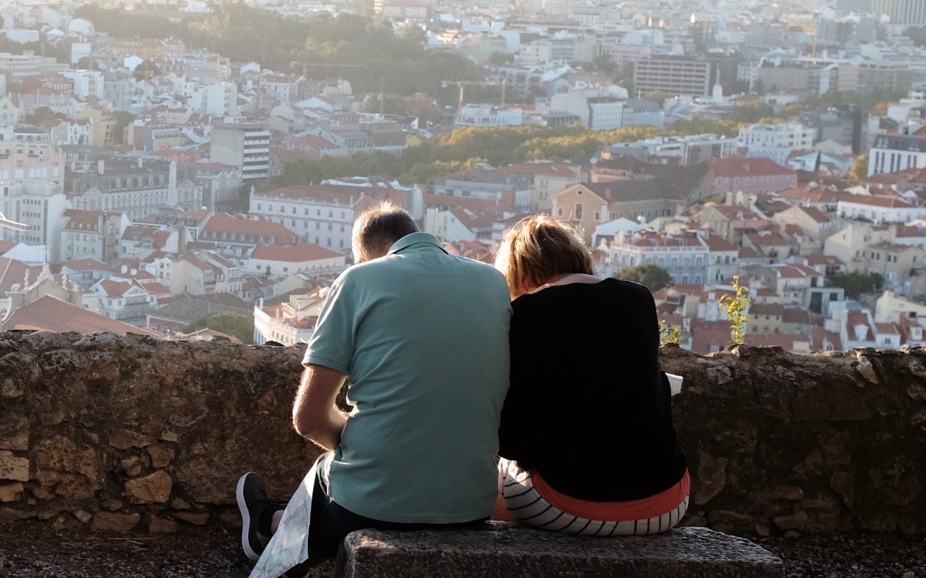 A couple sitting on a wall at a Lisbon viewpoint looking out over the city