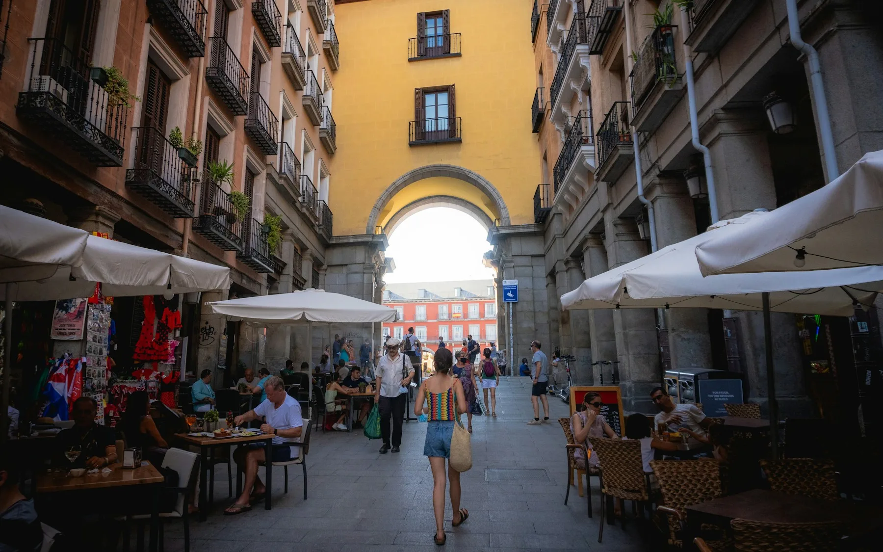 Two people walking through the arcades of the Plaza Mayor in Madrid, past café tables