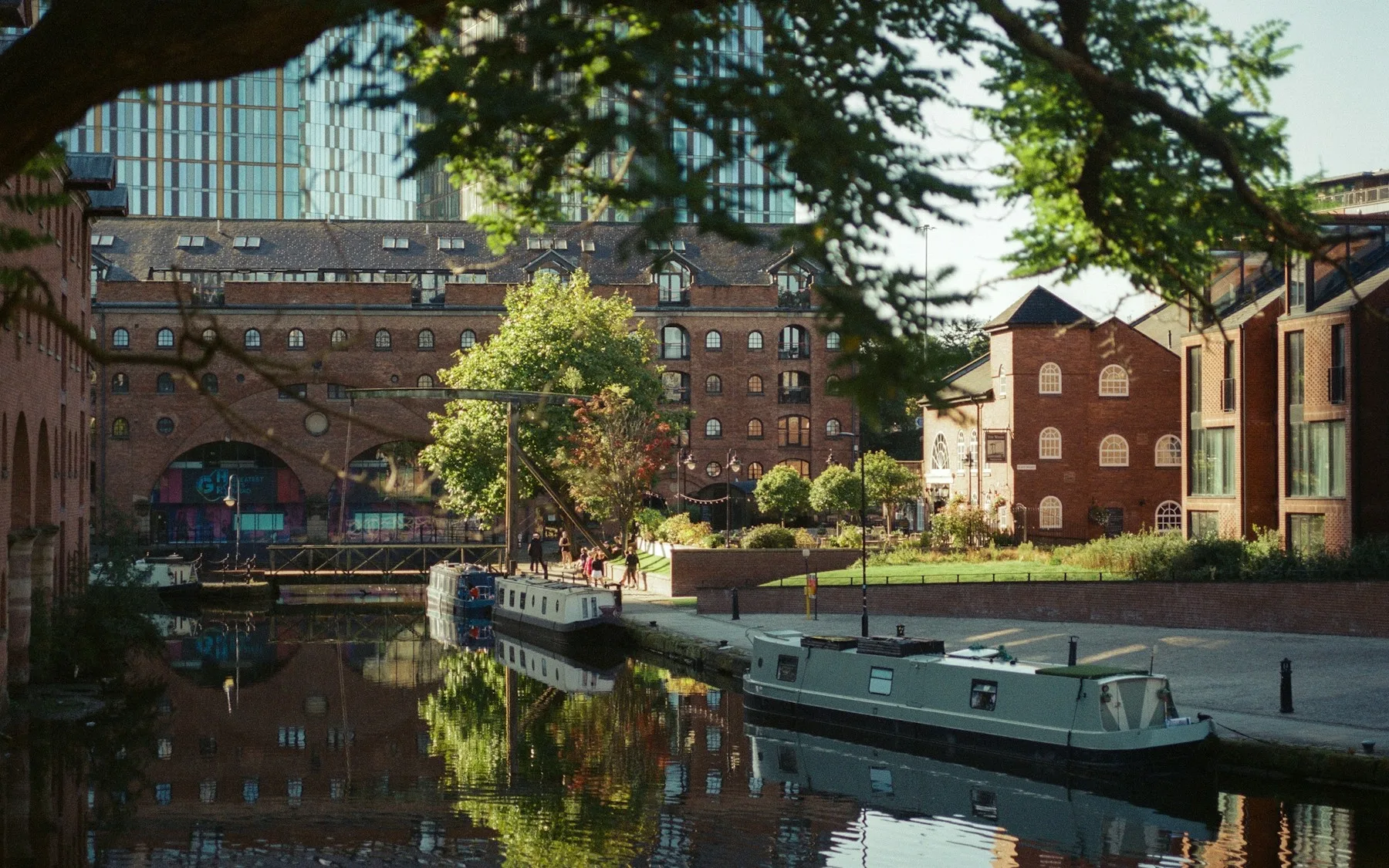 Narrow boats moored on a Manchester canal beside red-brick warehouses, with people on the towpath