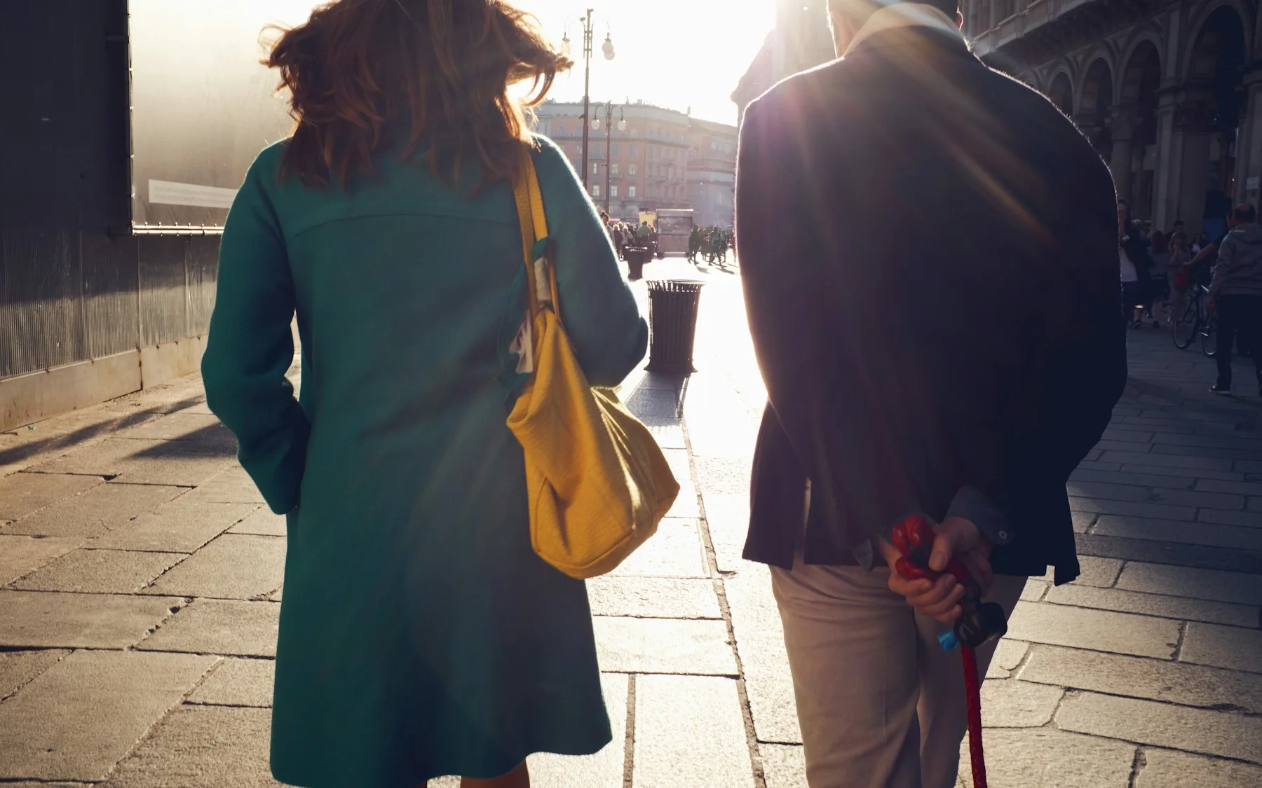 A woman and a man walking through a Milan street in low afternoon sun, photographed from behind