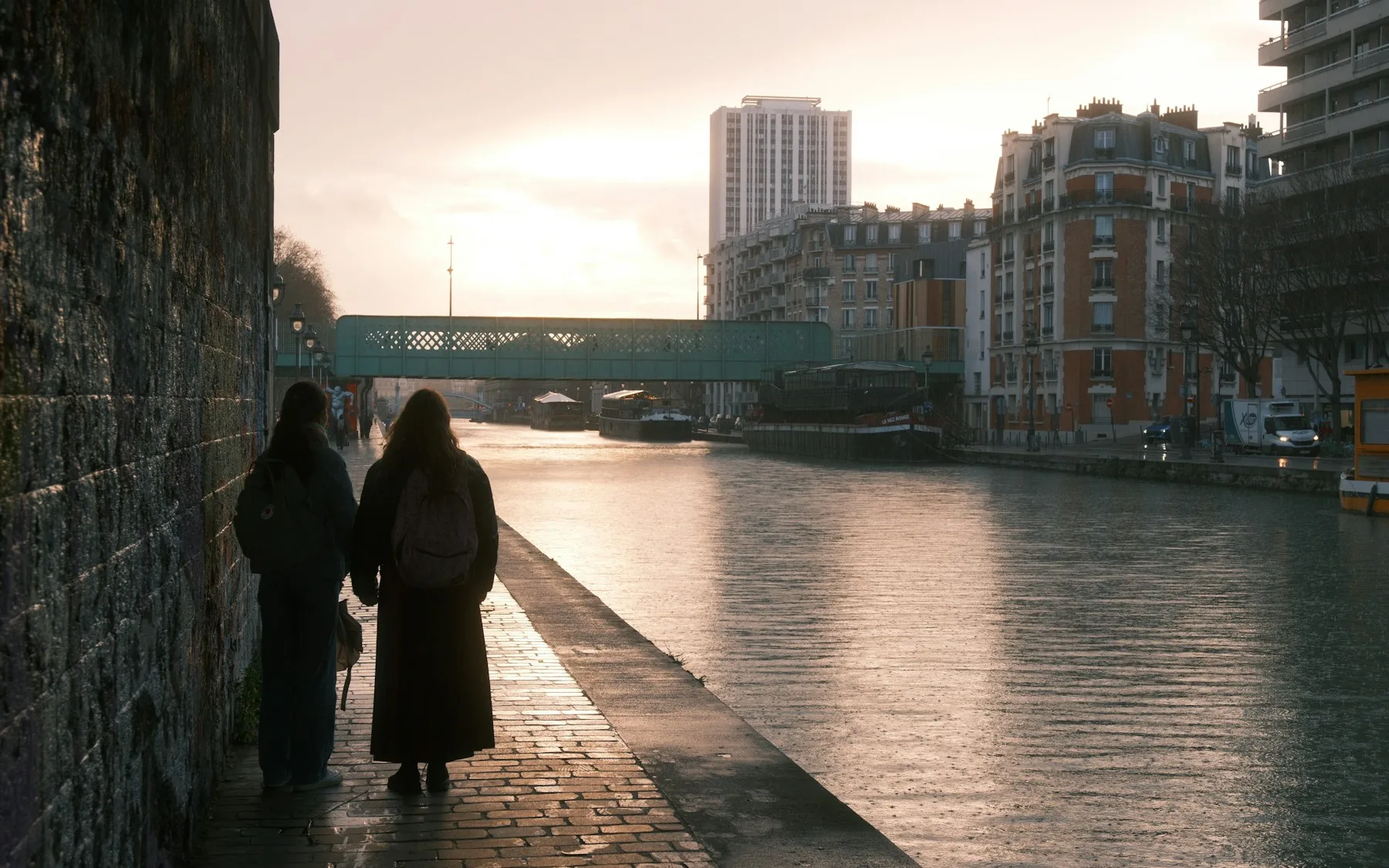 Two people walking along a Paris canal at sunset, photographed from behind