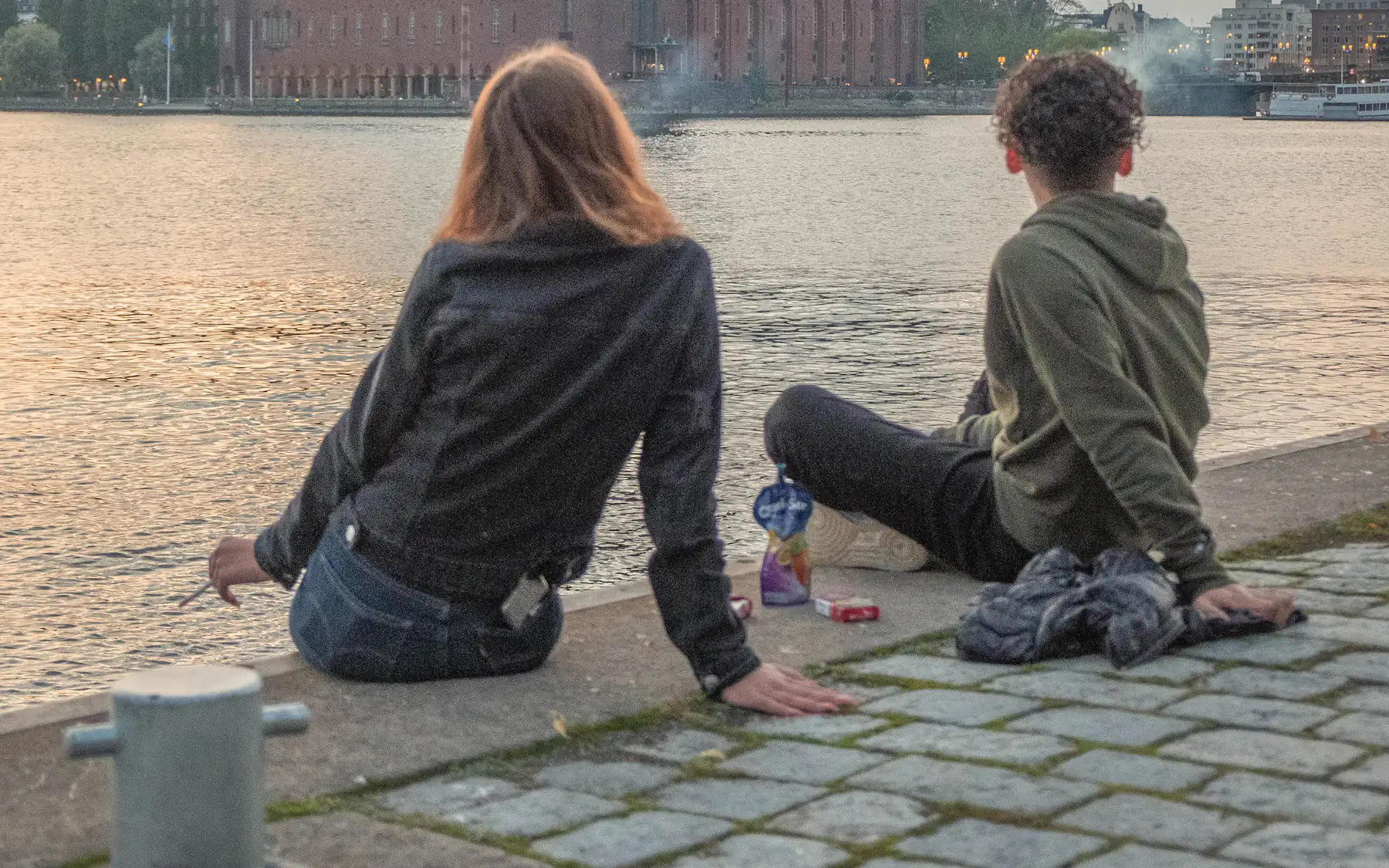 A couple sitting on a stone quay looking across the water at Stockholm City Hall